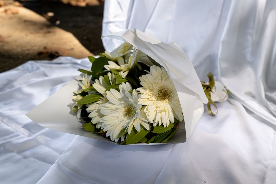 A bouquet of fresh white gerbera daisies and white chrysanthemums with green foliage, wrapped in white paper and tied with a gold ribbon, resting on a white satin-like fabric. The bouquet is displayed