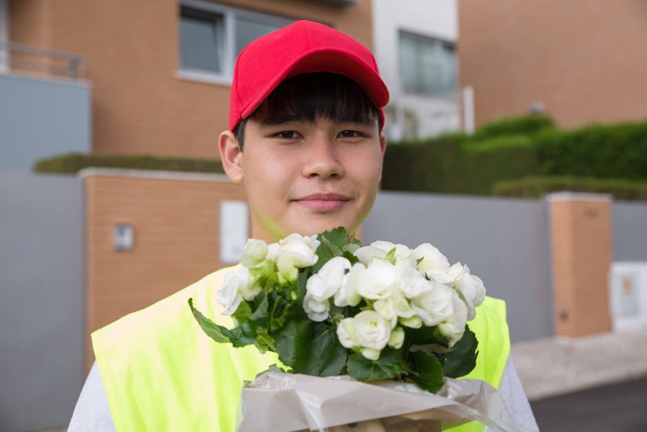 A young delivery person wearing a red cap and neon yellow safety vest holds a bouquet of fresh white roses with green foliage, wrapped in clear and white floral paper. The bouquet is prominent in the 