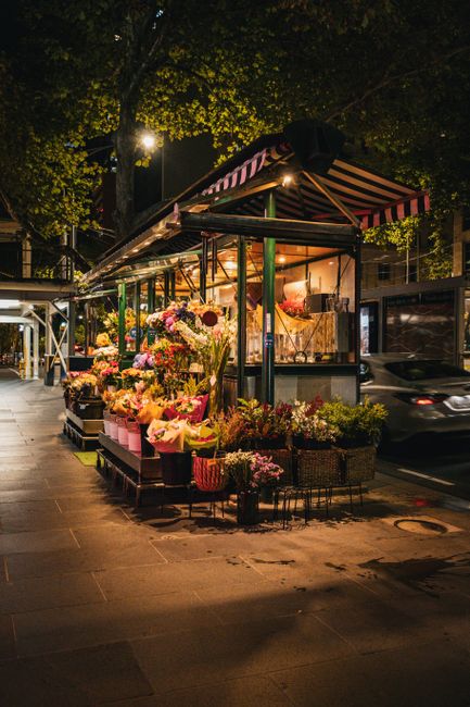 A street-side flower stall illuminated at night, displaying an array of fresh, vibrant floral arrangements including roses, lilies, and mixed seasonal blooms wrapped in pink and brown paper. The flowe
