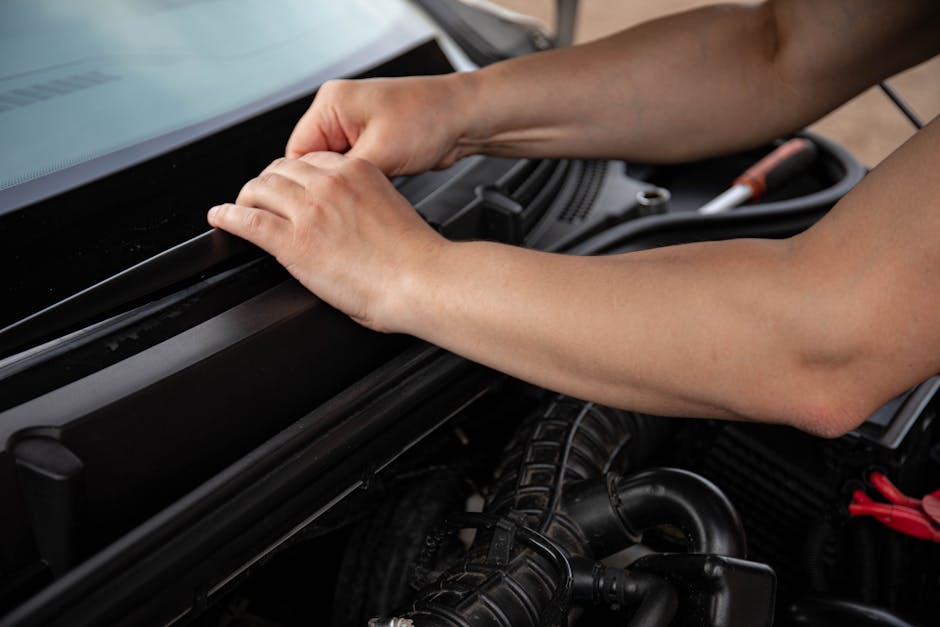 Close-up of a person's hands inspecting or repairing the engine compartment of a vehicle, with visible engine components, hoses, and tools in the background. The person’s arms are bare, focusing on 