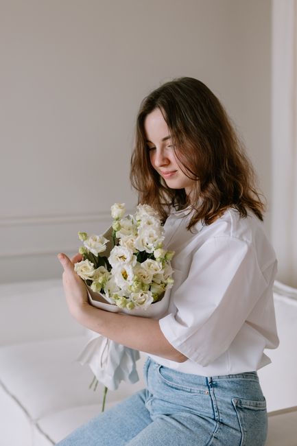 A young woman with shoulder-length brown hair, wearing a white shirt and light blue jeans, holds a bouquet of fresh white flowers with green accents, which appear to be lisianthus and possibly roses, 