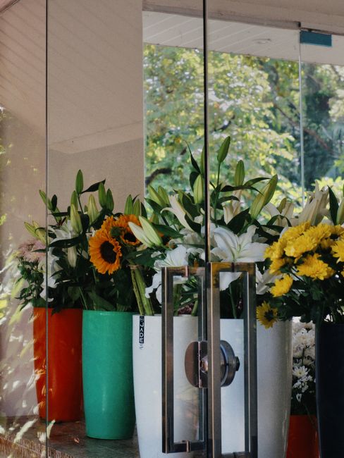 A collection of fresh floral arrangements displayed in various vases, including a white vase with white lilies and yellow chrysanthemums, an orange vase with sunflowers, and a turquoise vase with mixe