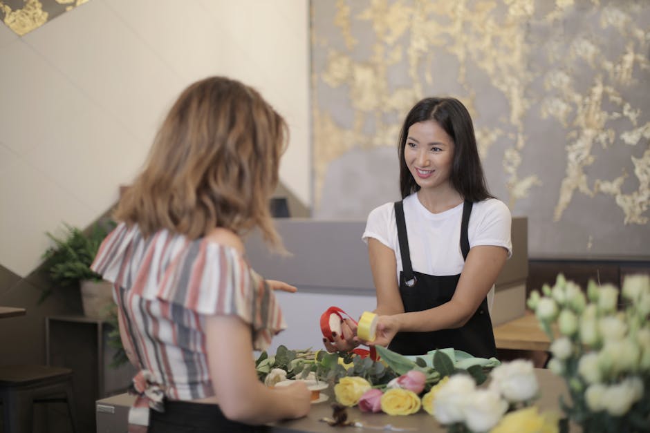 A young female florist with long dark hair, wearing a white t-shirt and black apron, is handing a floral gift wrapped with red ribbon to a customer with shoulder-length blonde hair in a flower shop. T