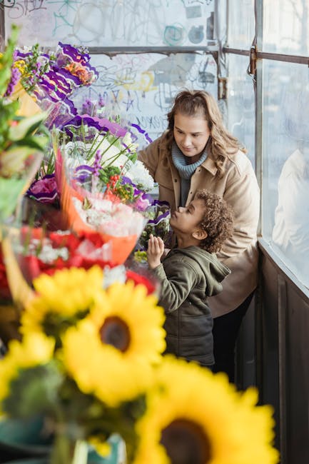 A woman and a young child are in a flower shop, surrounded by a vibrant display of fresh flowers including sunflowers, purple irises, and various mixed bouquets wrapped in colorful paper and cellophan